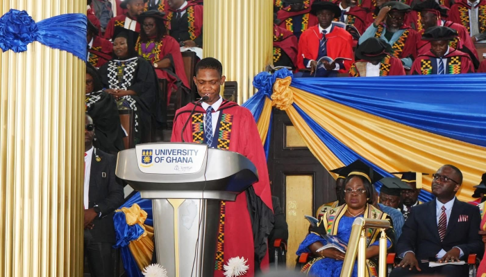 Worlanyo Tashie, wearing red and black academic robes, delivers the valedictory address from a University of Ghana lectern, surrounded by seated university officials and fellow graduates in the decorated Great Hall.