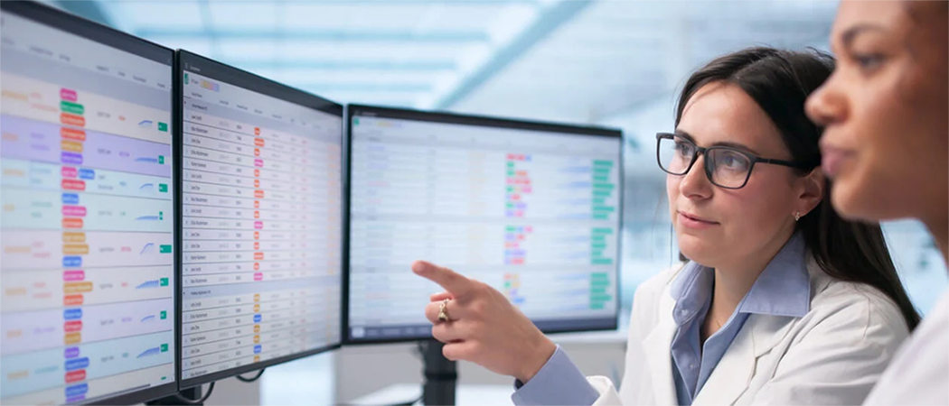Two healthcare professionals in white laboratory coats sit at a workstation in a modern hospital or clinical setting, reviewing medical data displayed across three large computer monitors.