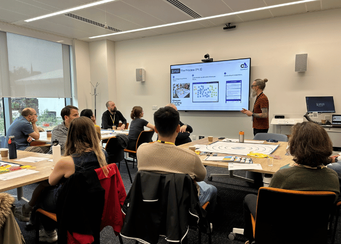 A group of people around tables with various objected lied out on them, a speaker in front of the screen at the back of the room