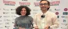 A woman and man stand looking into the camera hold two glass awards.