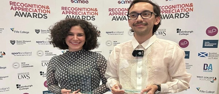 A woman and man stand looking into the camera hold two glass awards.