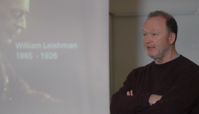 Professor Mike Barrett speaking beside a presentation slide showing Sir William Leishman and the dates 1865–1926.