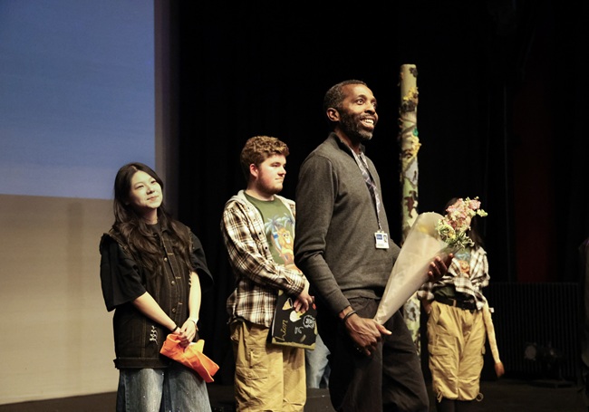 Dr Tawona Sitholé. Credit Abigail Morris A man holding a bunch of flowers stands in front of the cast of a play on stage