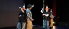 The main characters of a play Lion and Unicorn stand on a stage, two wear traffic cone hats as unicorns and two wear lion t-shirts