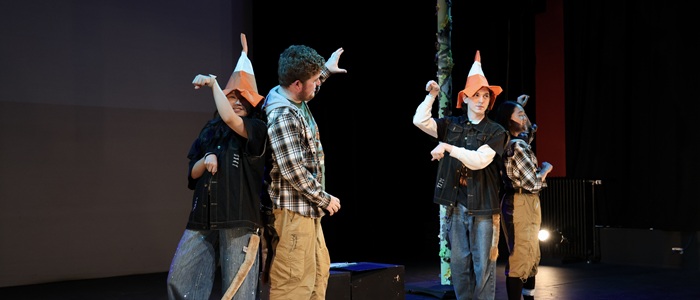 The main characters of a play Lion and Unicorn stand on a stage, two wear traffic cone hats as unicorns and two wear lion t-shirts