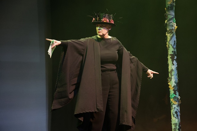 Susan Shaw as Cu sidhé. Credit Abigail Morris A woman wearing a black gown and hat stands on a stage for the Lion and Unicorn play