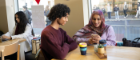 Two students sitting at a cafe table, one student is looking at their phone