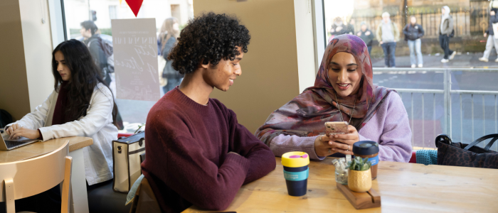 Two students sitting at a cafe table, one student is looking at their phone
