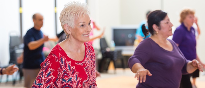 A light exercise class featuring a smiling woman with a red top and short white hair, and a woman wearing a purple top with a dark ponytail
