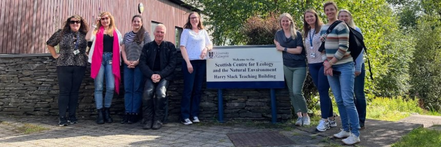 A group of School of Infection & Immunity colleagues stand outside the University of Glasgow’s Scottish Centre for Ecology and the Natural Environment. Director of Education Professor Nicola Veitch stands at the centre in a white t-shirt, with her hand on the sign.
