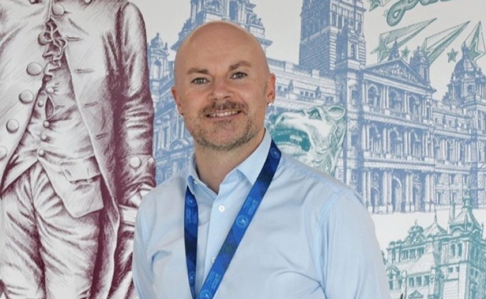 Kenny Logue standing in front of a wall mural in the atrium of the Adam Smith Building, University of Glasgow
