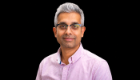 A head and shoulders portrait of Dr Rahul Kalla wearing glasses and a pink striped shirt against a black background.