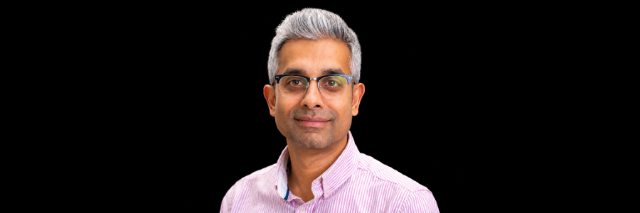 A head and shoulders portrait of Dr Rahul Kalla wearing glasses and a pink striped shirt against a black background.