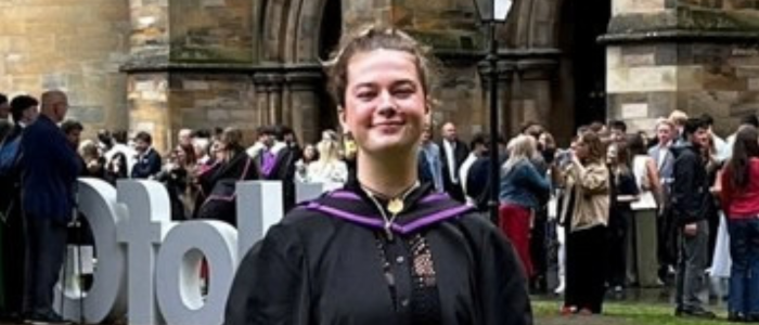 A young woman wearing a graduation gown smiling at the camera with people behind her
