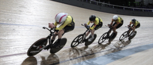 Two cyclists race around an indoor velodrome track, leaning into a bend on their bikes.