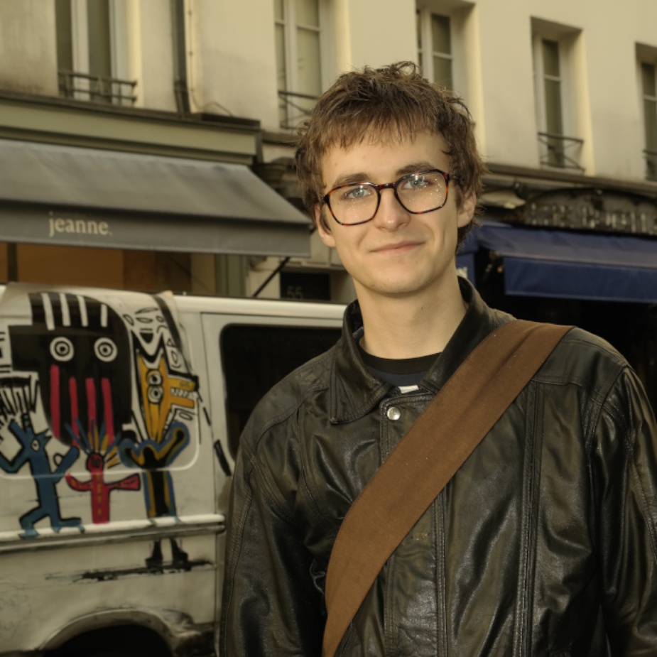 Sasha Diachenko stands in an urban streetscape. A painted van with colourful artistic designs is visible behind him, with shops and multi-storey buildings forming the background.