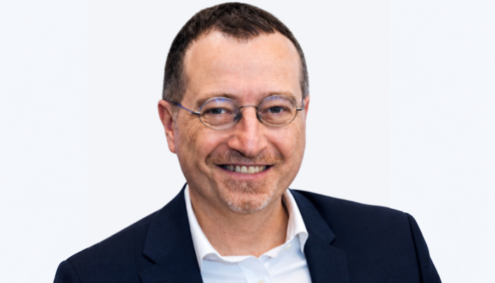 Professor Pasquale Maffia smiling at the camera, wearing glasses, a navy jacket and a white shirt against a pale grey background.