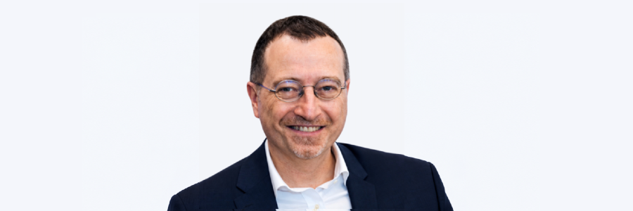 Professor Pasquale Maffia smiling at the camera, wearing glasses, a navy jacket and a white shirt against a pale grey background.