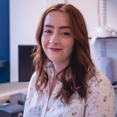 Dr Sarah K Walsh of the MRC-University of Glasgow Centre for Virus Research (CVR), smiling at the camera in a light-coloured floral blouse, with shoulder-length auburn hair.