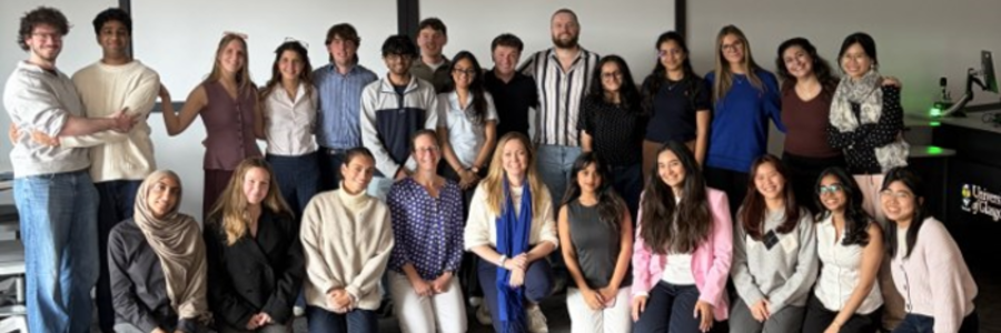 Students and staff from the Business in the Biosciences course pose in two rows in a teaching room. Course leads Dr Anna McGregor, wearing a patterned blue blouse, and Dr Laura McCaughey, wearing a blue scarf, kneel at the centre of the front row.
