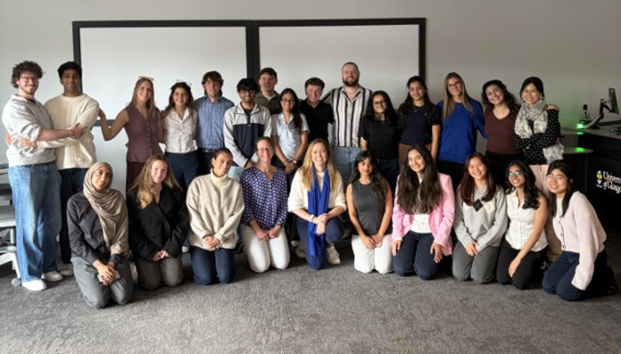 Students and staff from the Business in the Biosciences course pose in two rows in a teaching room. Course leads Dr Anna McGregor, wearing a patterned blue blouse, and Dr Laura McCaughey, wearing a blue scarf, kneel at the centre of the front row.