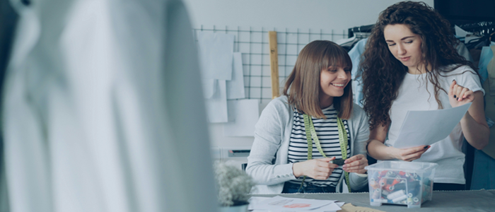 Two women in a workshop, standing behind a work desk reading a sheet of paper.