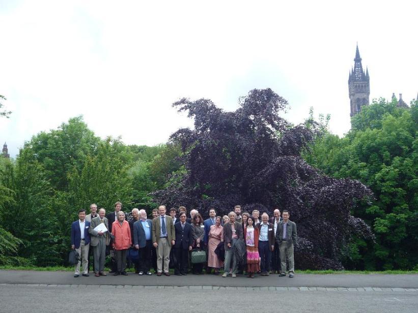 photograph of exterior: a big group of older people of mixed gender and ethnicity, in formal or business attire, posing in front of lush greenery with the tower of university of glasgow peeking in the background