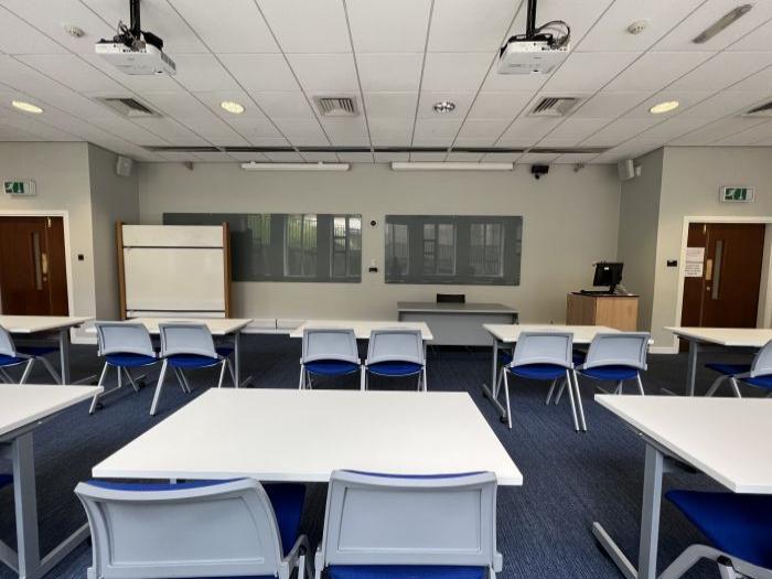 Flat floored teaching room with rows of tables and chairs, glassboards, moveable whiteboard, projectors, PC, and lecturer's table and chair.