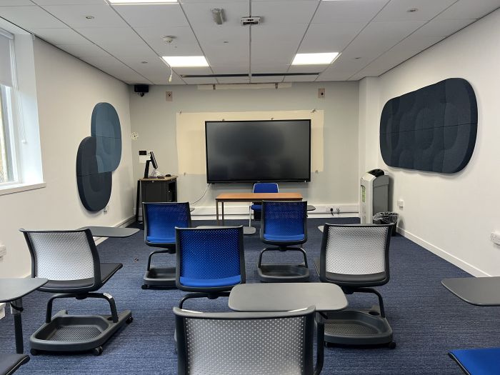 Flat floored teaching room with tables and chairs in round table set-up, projector, whiteboards, screen, and PC