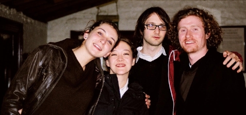 photograph of interior: a group of four young white students huddled together, smiling at the camera. background is evening outside