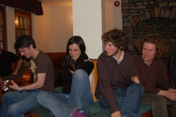 photograph of interior: four young students (three men and a woman) in jeans and jumpers, sitting on a bench relaxing. one man has a guitar which he is playing. The woman is smiling embarrased, while the man to her rigth is looking attentively at her