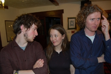 photograph of interior: three white young students (a woman and two men) in a room. The woman (in the middle) is talking to the man to her right, while the otehr man with curly blond hair seems to be talking to someone out of the picture
