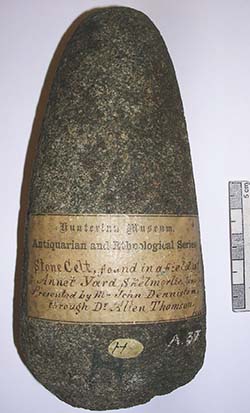 A Celtic Stone Axe head is shown in close-up. Across the front of the axe head there is an old sepia toned museum label describing the object as 