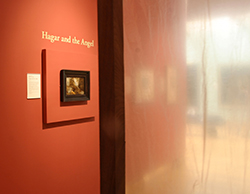 On the left hand side of the image, a small framed painting hangs on a rust colured gallery wall but is shown at an angle and cannot be seen in detail. A dark strip cuts the picture in half - this is the wooden post at the corner of the sound and sculpture installation.  To the right of the post, the image is blurry as we are viewing the gallery through a translucent plastic sheet.