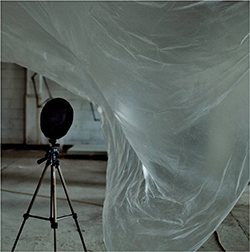 photograph of a modern sculpture accompanying the artists sound installation. There is an instrument on a tripod in the left hand corner.  The rest of the image is of a sheet of translucent plastic caught in a moment of movement that suggest flapping caused by air motion.  imagine the sound the plastic would make