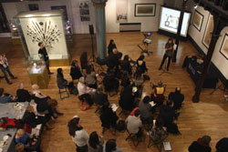 a museum event. A speaker stands at a microphone and about 30 people are seated watching