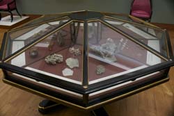 a photograph of a wooden museum cabinet. we can see 4 of the 8 sides of the glass topped cabinet. there are objects inside the case, apparently rocks and shells