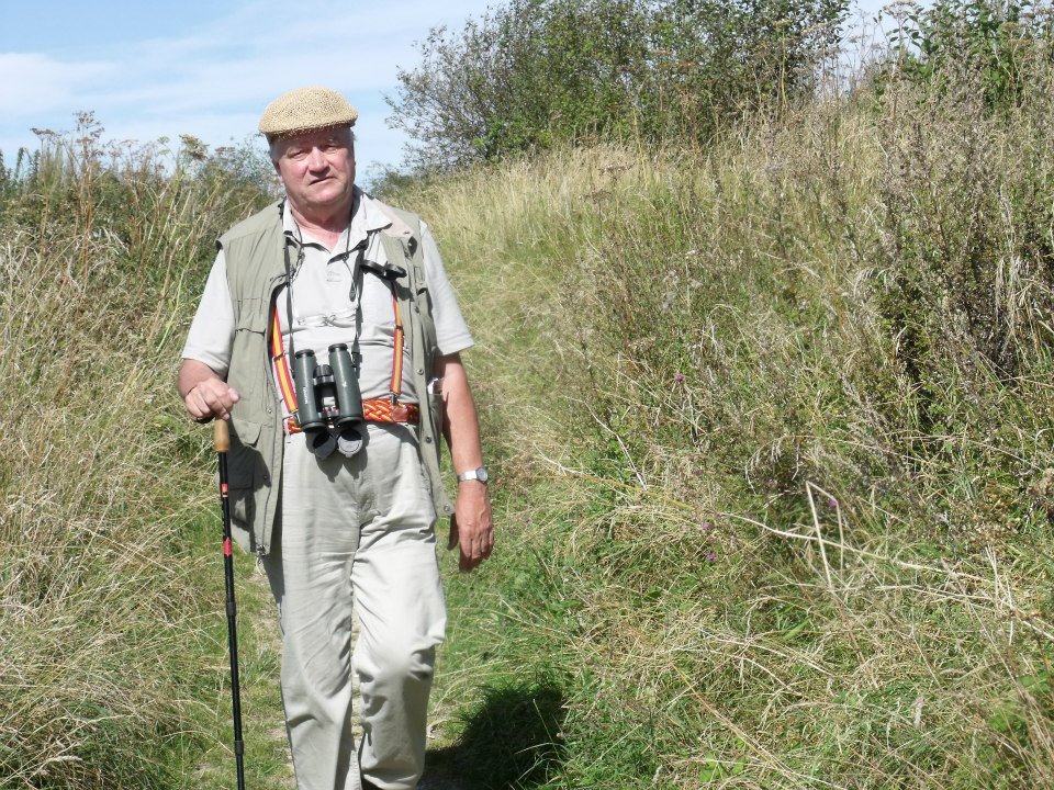 photo of Prof Dudley Knowles walking in the a lush green environment, dressed in beige and white, with a flat cap, using a walking stick