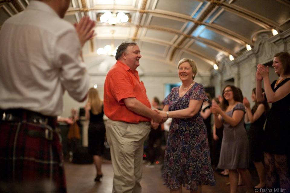 photograph of Prof Dudley Knowles at his retirement ceilidh in a beautifully vault-ceiling building, holding hands with a middle-aged white woman in a dress, surrpunded by other onlookers clapping