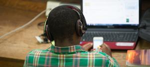 Student facing computer screen wearing headphone while looking at phone with study book beside him.