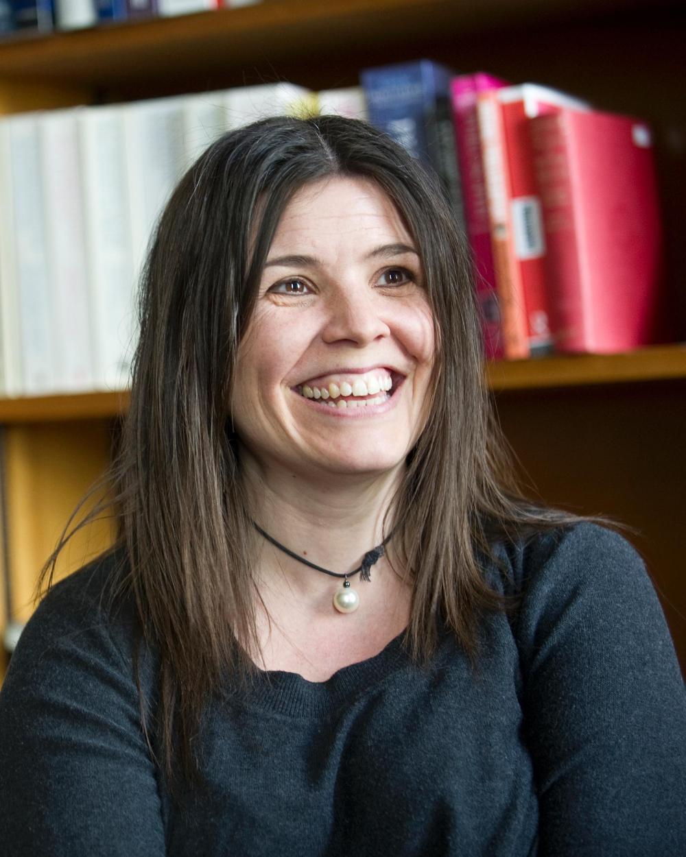 photo of Fiona Macpherson: headshot of a younger white woman with long brown hair, a black choker and black blouse, smiling against the backdrop of a full bookshelf