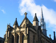 photograph of a historical ecclesiastical architecture with big volumes, small spires and a cross on top. in the background, the white spire of the University of Glasgow tower, against a bright blue sky