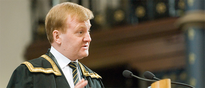 photo of Charles Kennedy: old man with yellow hair in university regalia with golden embroidered shoulders, wearing a tie and white shirt underneath, speaking in two long microphones