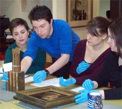 3 students, one standing, two sitting, all wearing blue gloves and handling and condition checking museum objects. Hunterian Print Room.