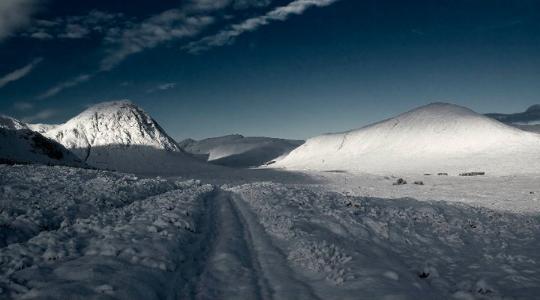 photograph of outdoors: Snowy landscape with tracks going ahead of the camera, two sun-lit hills completely covered in snow in the background