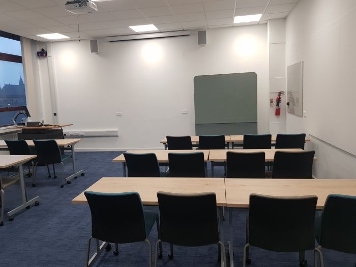 Flat floored teaching room with rows of tables and chairs, movable glassboard, projector, screen, PC  and lectern.