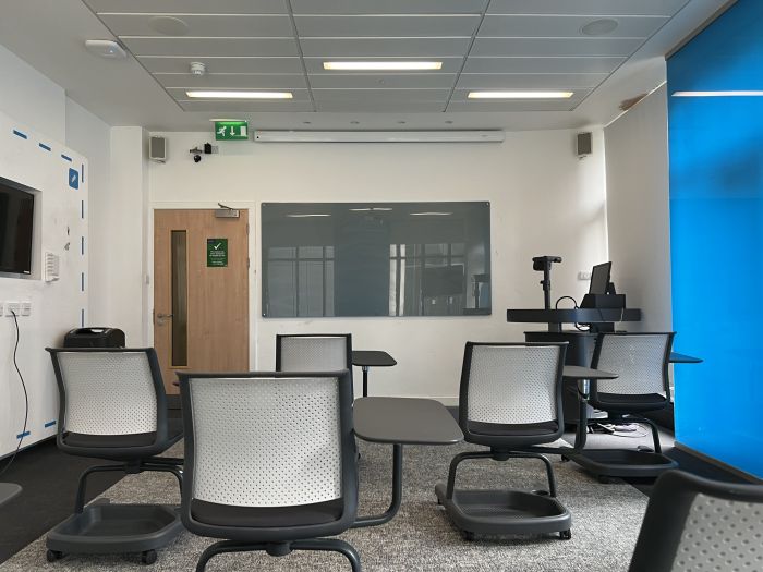 Flat floored teaching room with tablet chairs, large monitor, visualiser, PC, lectern, and glassboard.