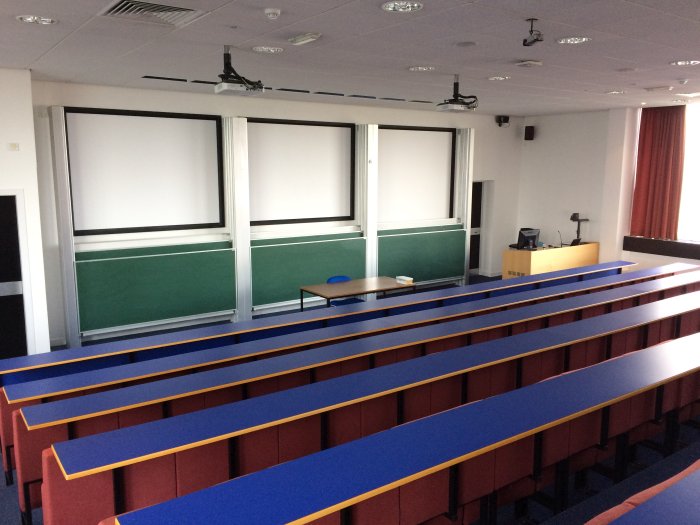 Raked lecture theatre with fixed seating, projectors, screens, chalkboards, visualiser, and PC