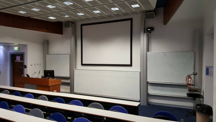 Raked lecture theatre with fixed seating, screen, whiteboards, visualiser, and PC
