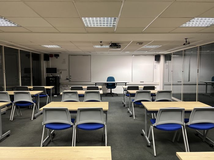 Flat floored teaching room with rows of tables and chairs, lecturer's table and chair, whiteboards, projector, and PC.